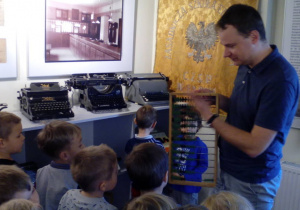 Kustosz demonstruje dzieciom najstarszy przedmiot do liczenia - liczydło. W tle maszyny do pisania i liczenia.